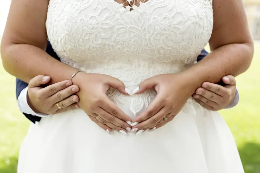 Detal białej sukni ślubnej plus size z dłońmi ułożonymi w serce na brzuchu kobiety
